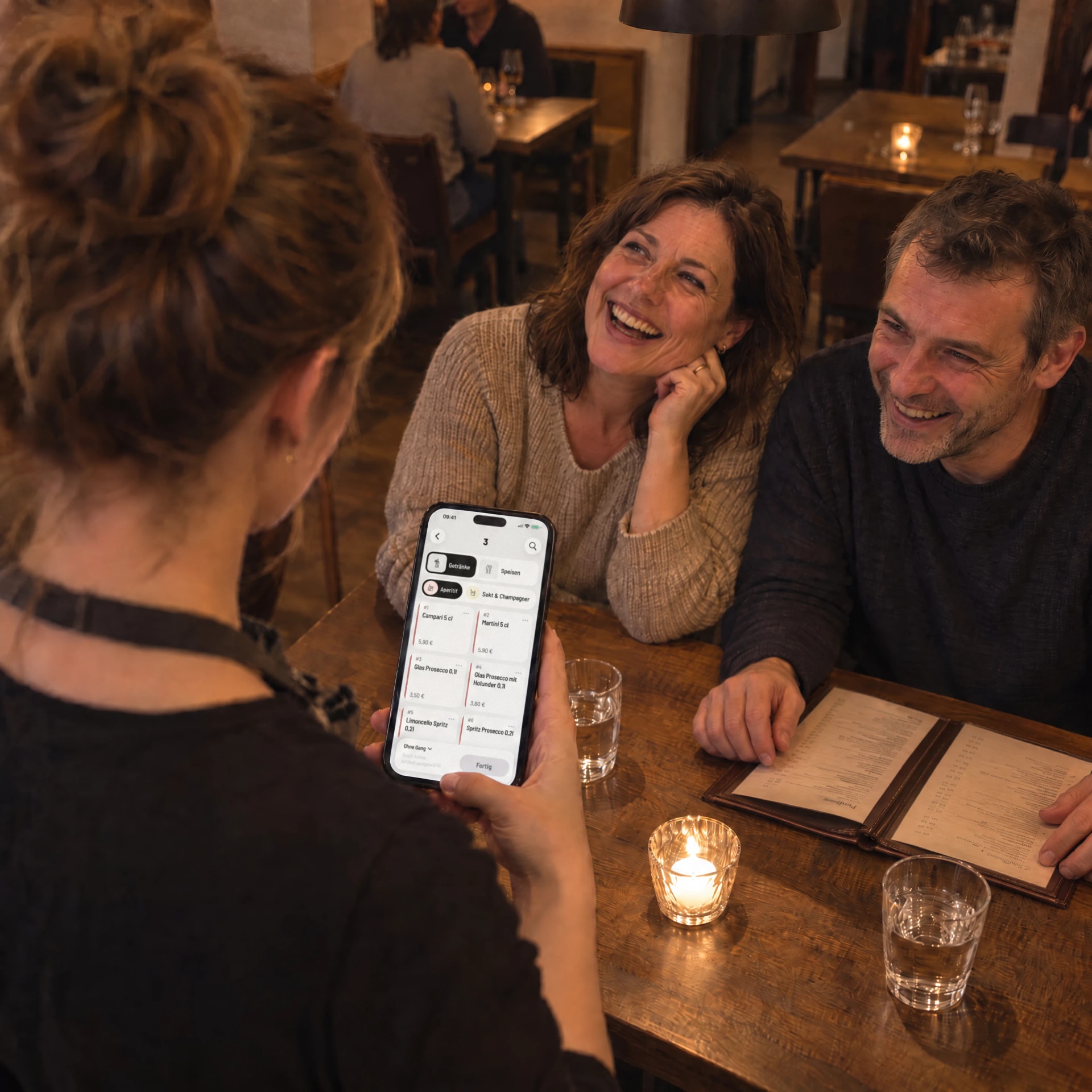 Servicekraft nimmt mit der Servire-App auf dem iPhone die Getränkebestellung eines Paares auf, beide lachen