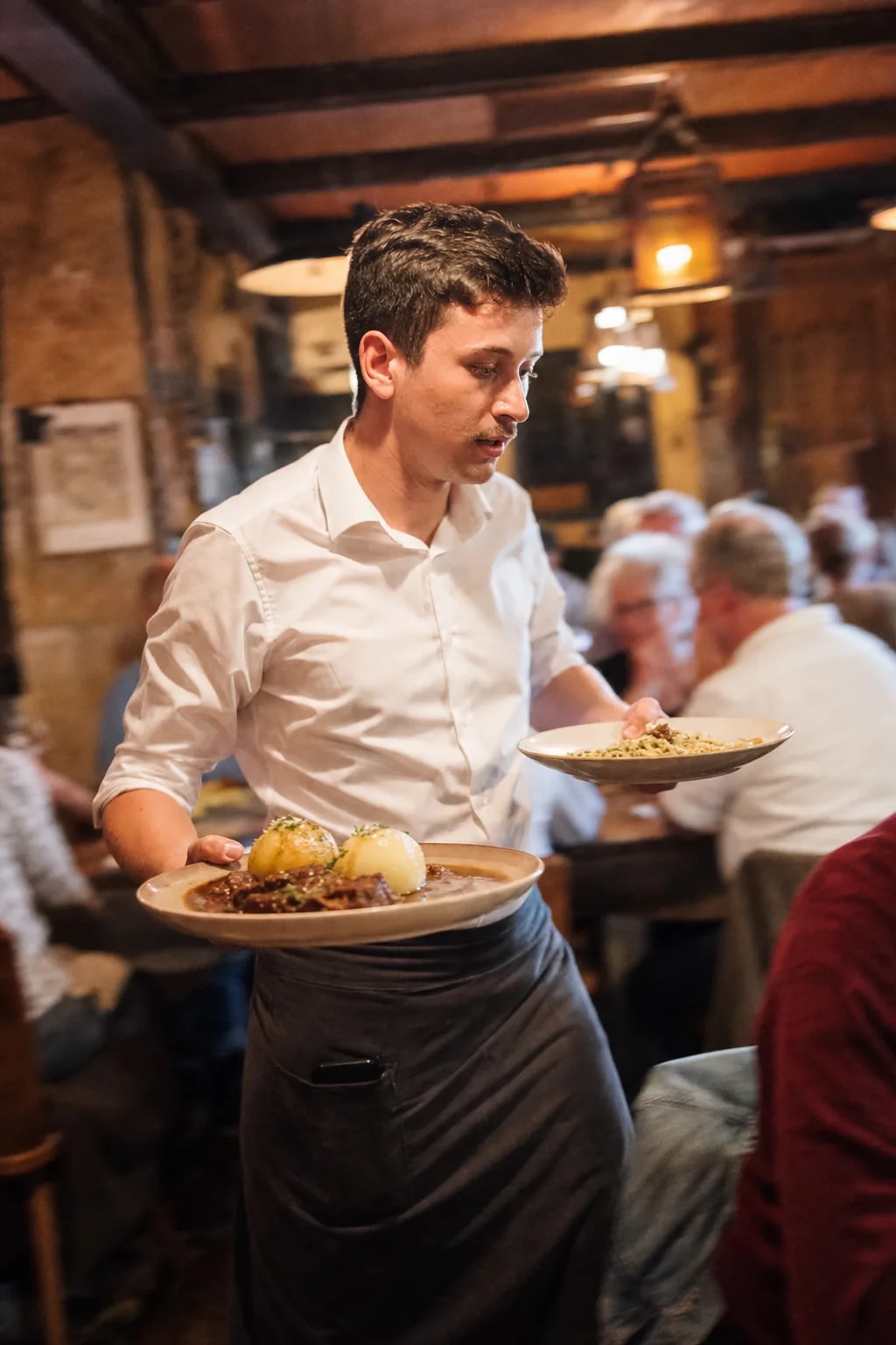 Waiter carrying plates through the busy dining room, his phone tucked in his apron pocket