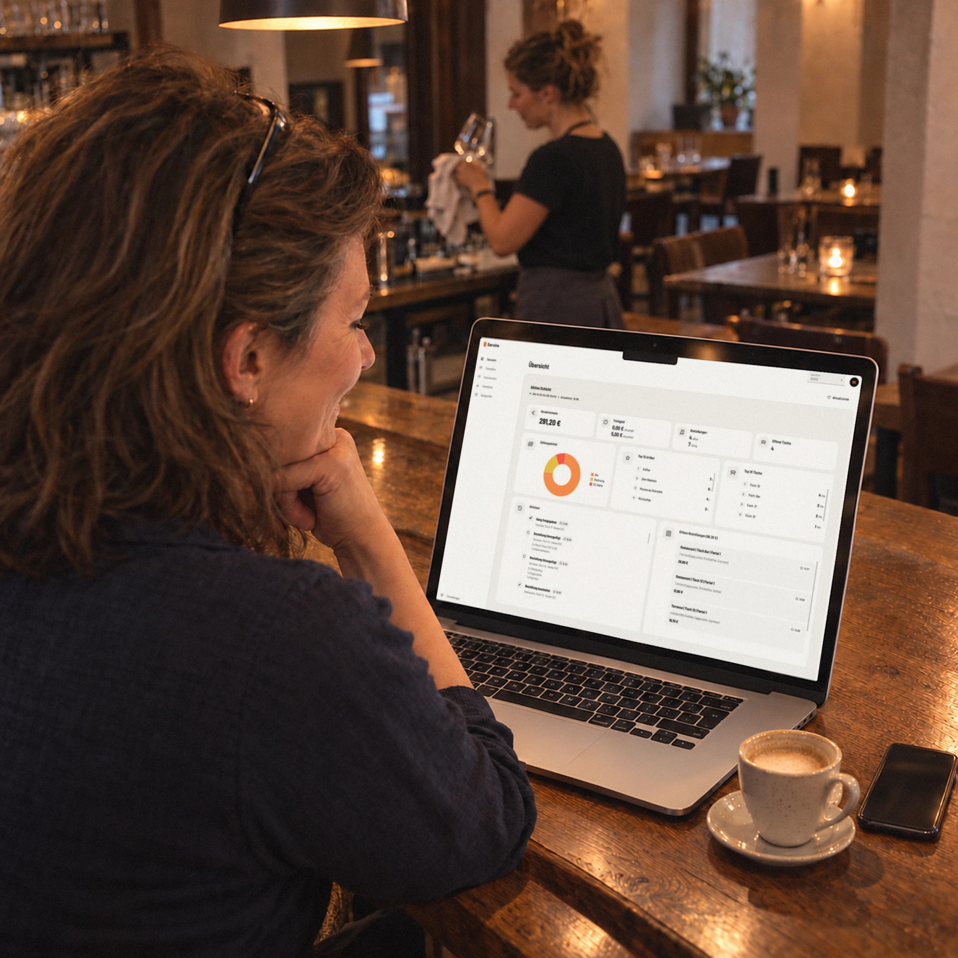 Die Wirtin sitzt vor der Öffnung an der Theke und sieht im Web desk auf dem Laptop die Übersicht des Tages